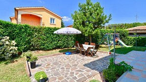 A patio with stone flooring, a dining table with chairs, a white umbrella, and a playground with a slide. - Relaxing rural apartment with playground and grill (Nedescina)