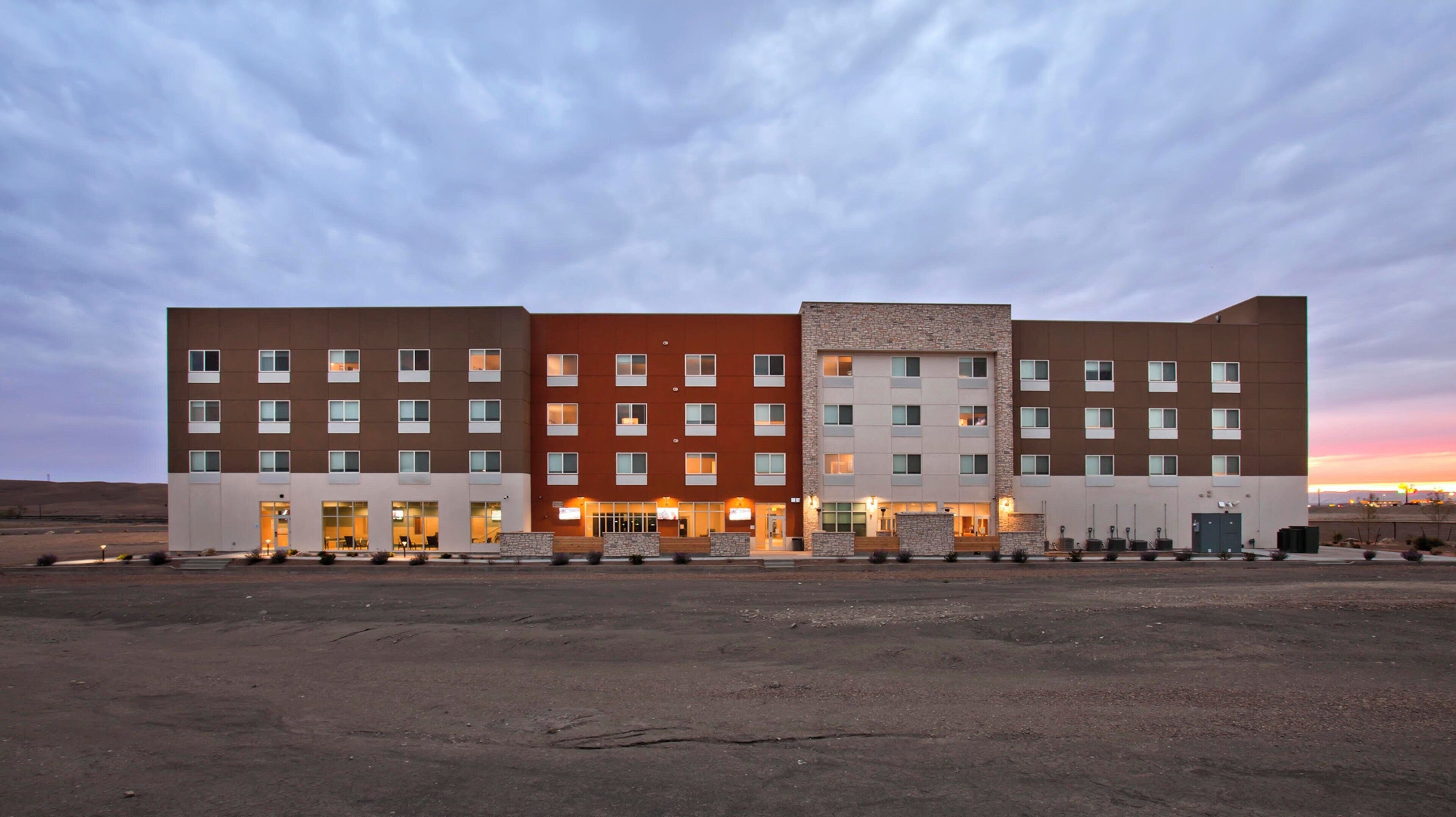 Um hotel com vários andares, telhado plano, entrada central e estacionamento em frente.