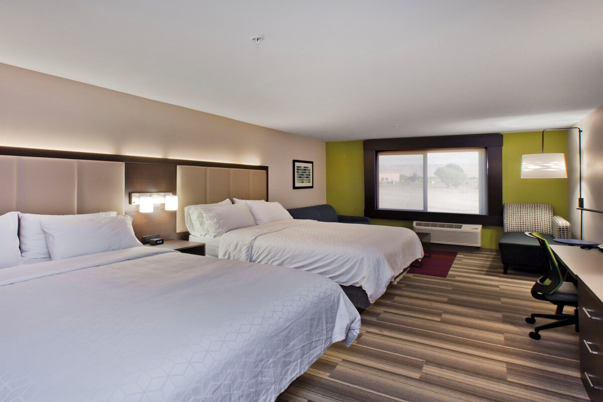 A hotel room with two beds, a desk, a chair, a window, and a wall-mounted air conditioner.