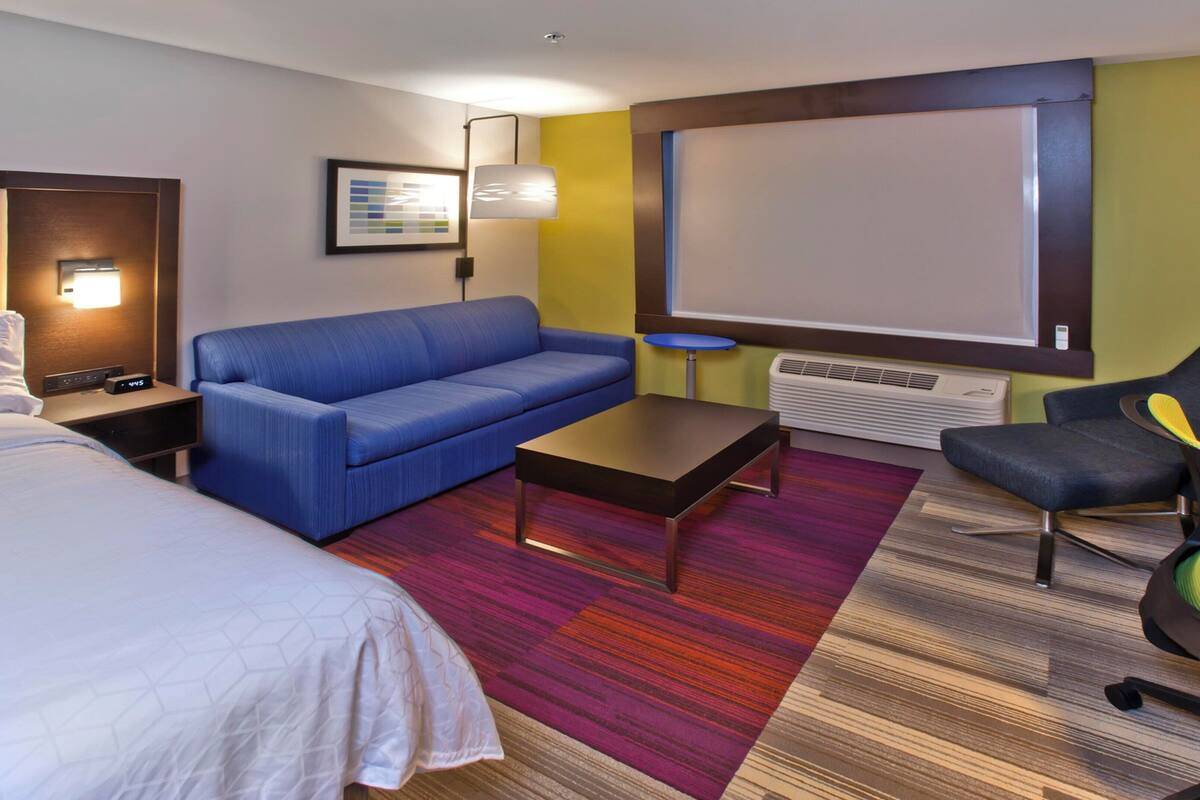 Une chambre d’hôtel avec un lit, un canapé bleu, une petite table, un bureau avec une chaise, un lampadaire, un tableau encadré et une grande fenêtre.