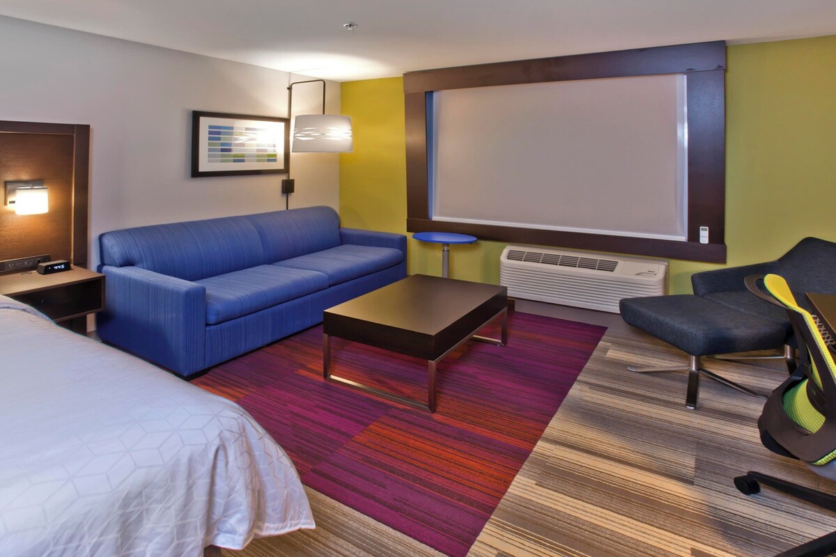 A hotel room with a bed, a blue sofa, a small table, a desk with a chair, a standing lamp, a framed picture, and a large window.