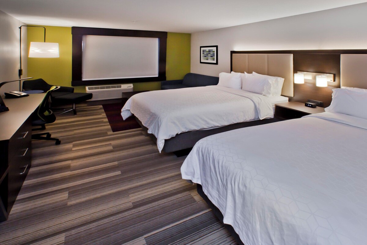 A hotel room with two beds, a desk, a chair, a lamp, and a large window.