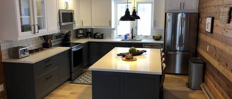 Una cocina moderna con armarios blancos, una isla central, electrodomésticos de acero inoxidable e iluminación colgante.