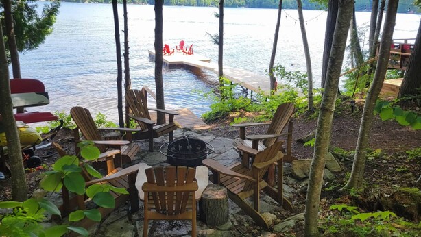 Une terrasse au bord du lac, avec des chaises en bois et un foyer, entourée d’arbres et offrant une vue sur l’eau.
