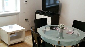 A small dining area with a round glass table, black chairs, a white side table, a black TV stand, and a clock on the wall.