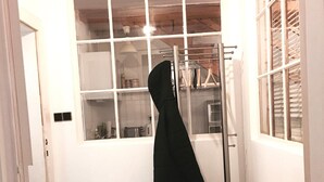 Ein kleiner, mit Fliesen ausgelegter Raum mit einem großen Fenster, einem schwarzen Mantel an einem Kleiderständer, einem Korbwäscheschrank und einem Paar Schuhe auf dem Boden.