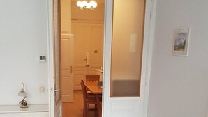 A room with a wooden herringbone floor, a white bench, a white dresser, and a door leading to a dining area with a chandelier.