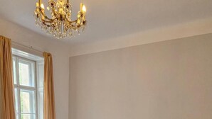 A bedroom with a large bed, a bedside table, a nightstand, a lamp, a window with curtains, and a chandelier.