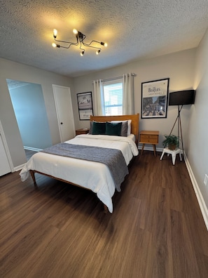 Un dormitorio con cama, mesita de noche, lámpara de pie, ventana con cortinas, puerta y un cartel en la pared.