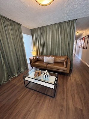 Una sala de estar con un sofá de cuero marrón, una mesa de centro de vidrio con adornos, cortinas verdes y un pasillo con cuadros enmarcados.