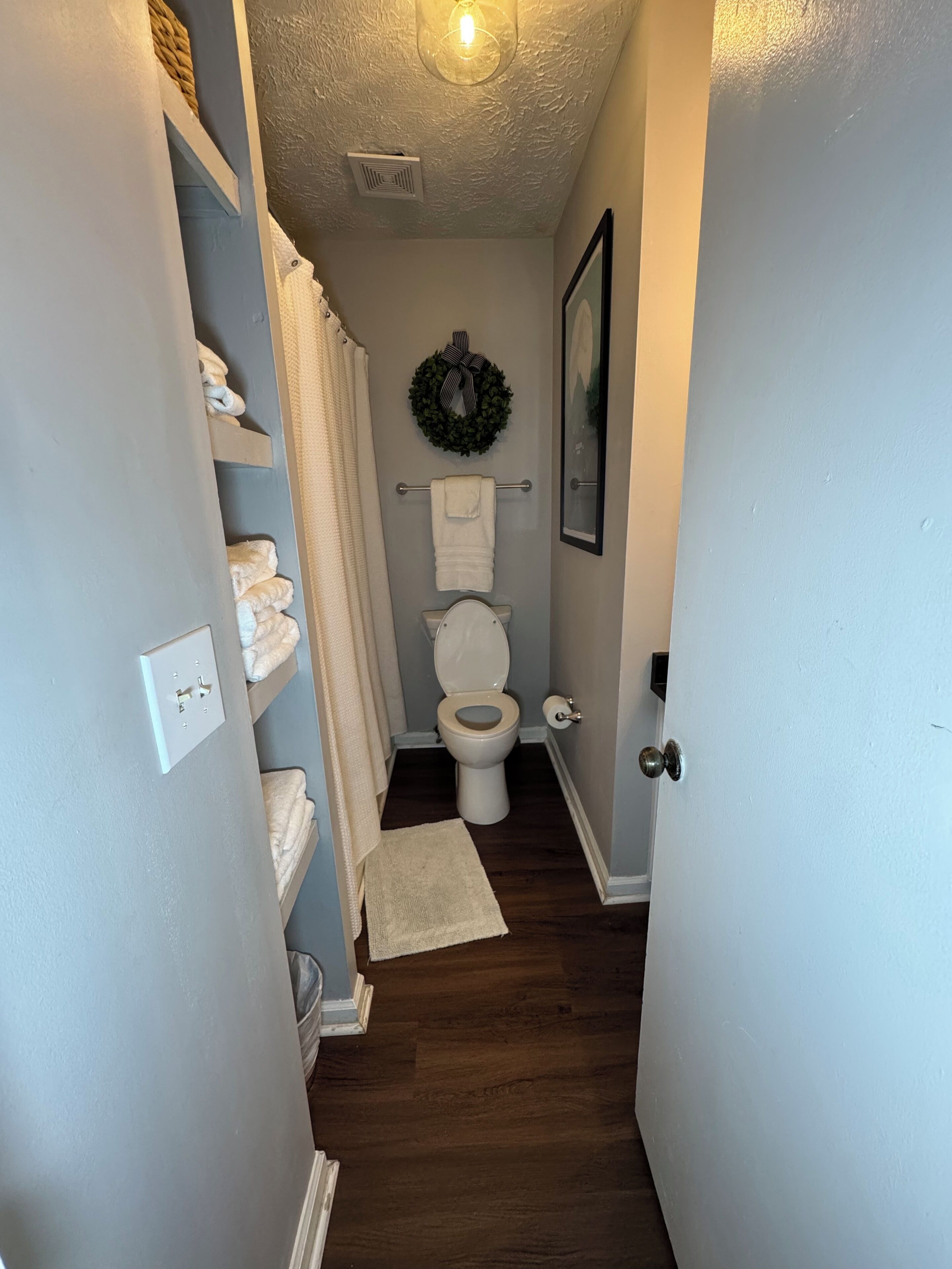 Un couloir étroit mène à une salle de bain équipée de toilettes, d’un porte-serviettes et d’une guirlande accrochée au mur.