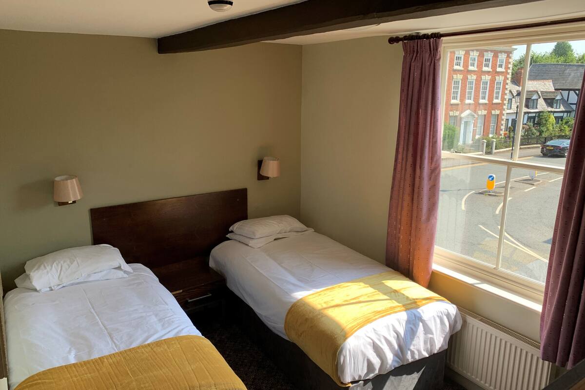 A hotel room with two single beds, a window with curtains, a radiator, and a view of the street outside.