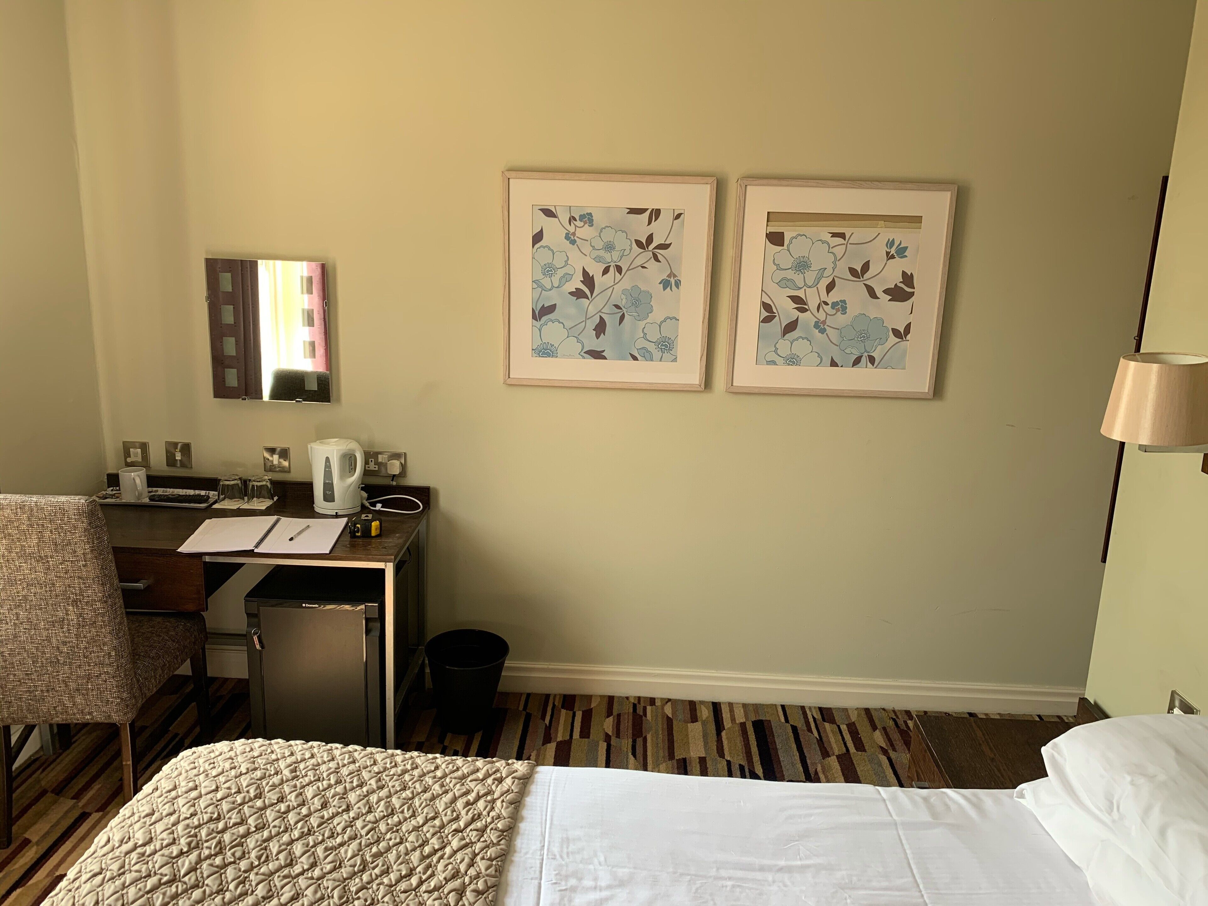 Habitación de hotel con cama, escritorio con hervidor, silla y dos cuadros con motivos florales en la pared.