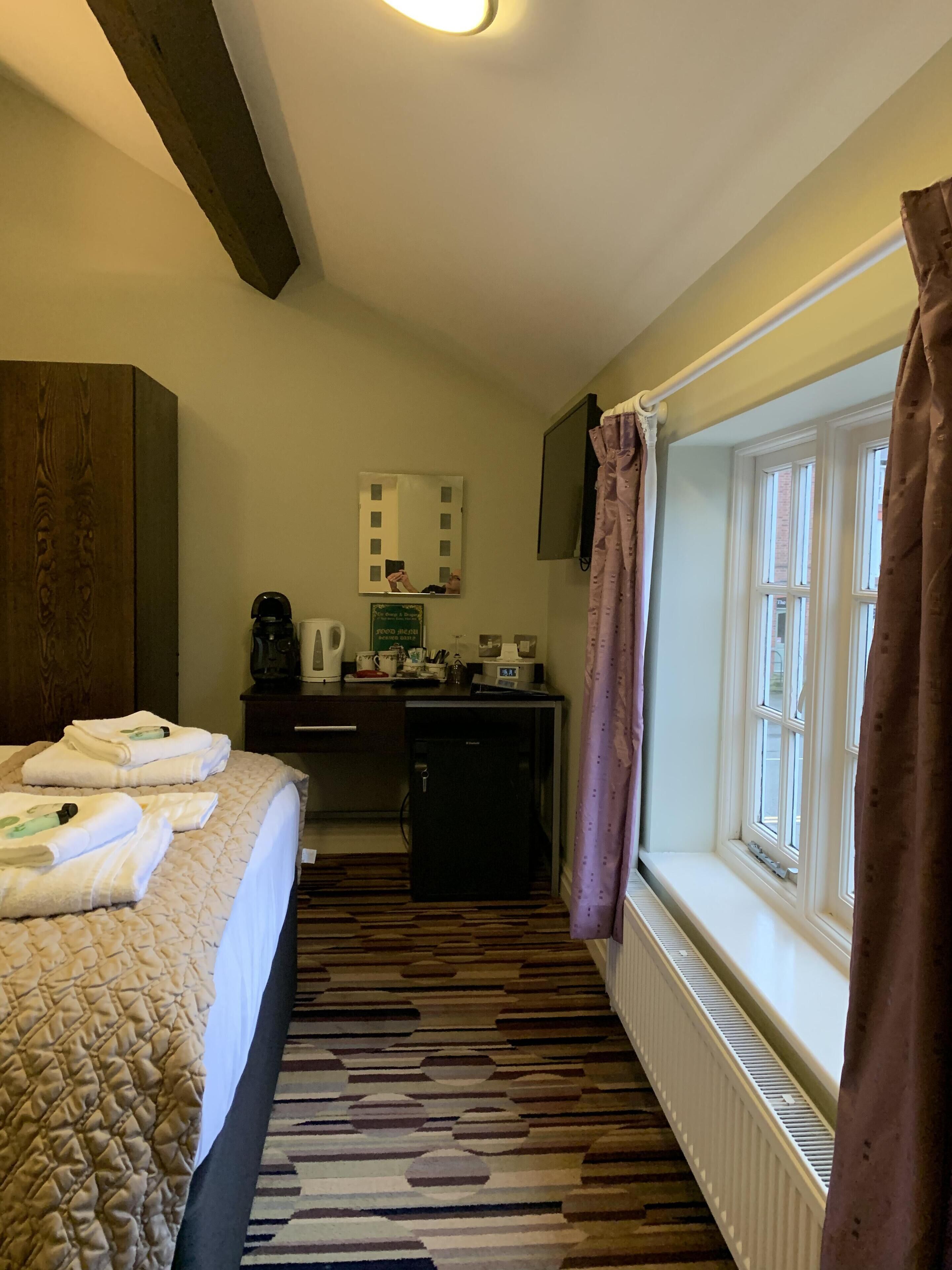 A hotel room with a bed, a small kitchenette, a mirror, and a window with curtains.