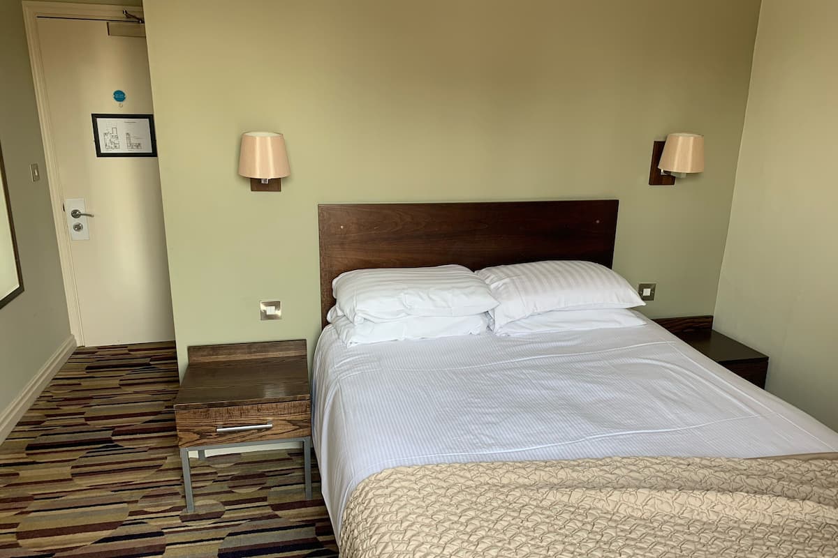 A hotel room with a wooden headboard, a bed with white linens, two bedside lamps, a patterned carpet, and a door with a light switch.