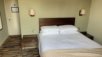 Habitación de hotel con cabecera de madera, cama con ropa blanca, dos lámparas de noche, alfombra estampada y una puerta con interruptor de luz.
