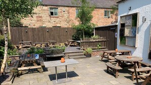 A patio area with wooden tables and benches, stone flooring, and a brick building in the background. - George & Dragon (Chester)