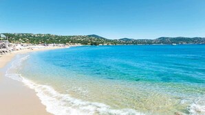 A beach with clear blue water, sandy shore, and a hilly landscape in the background.