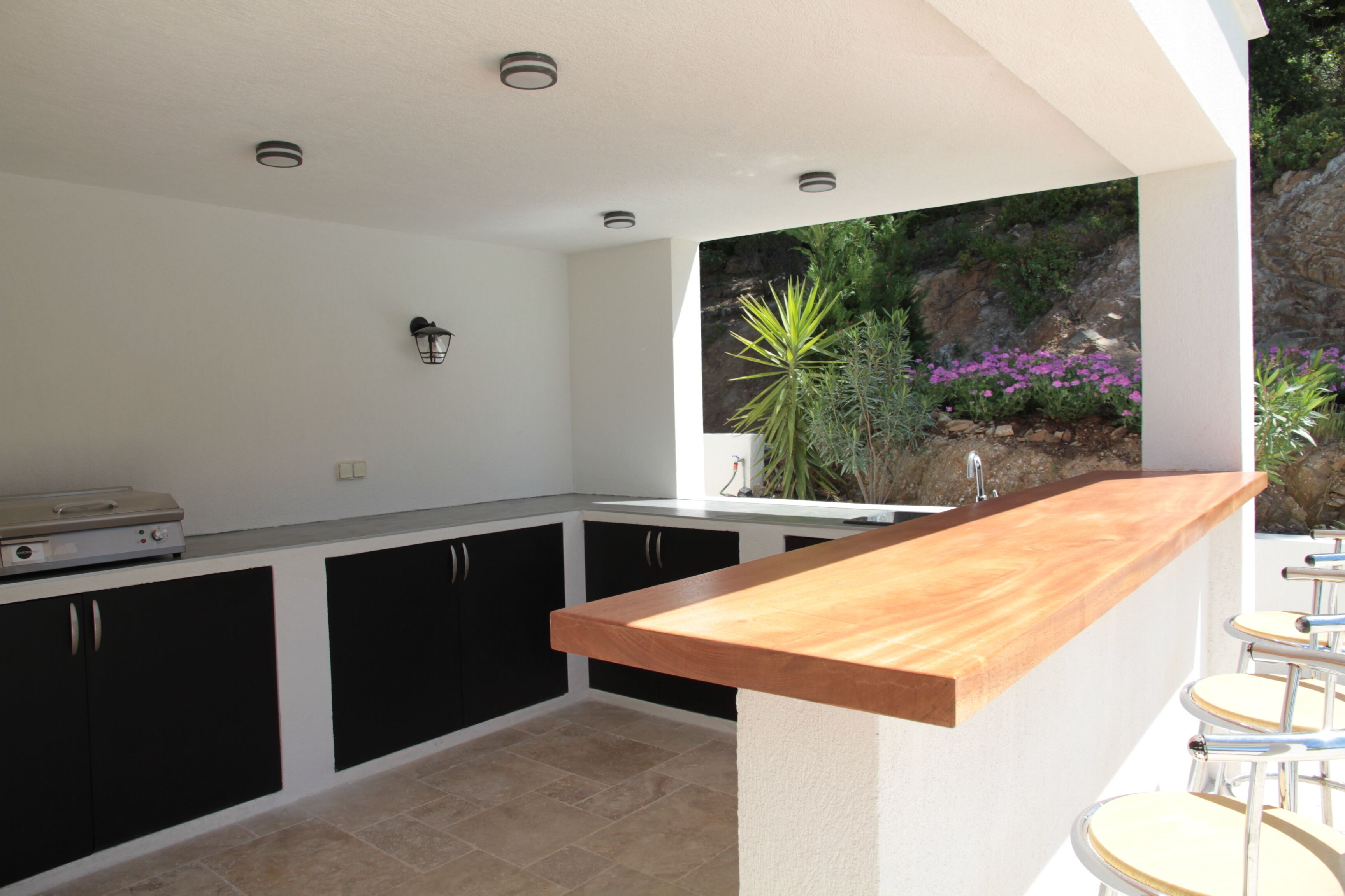 A modern kitchen with black cabinets, a wooden countertop, and a view of a rocky garden with plants and flowers.