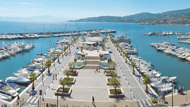 A marina with boats docked, a central walkway, and a view of the sea and distant hills.