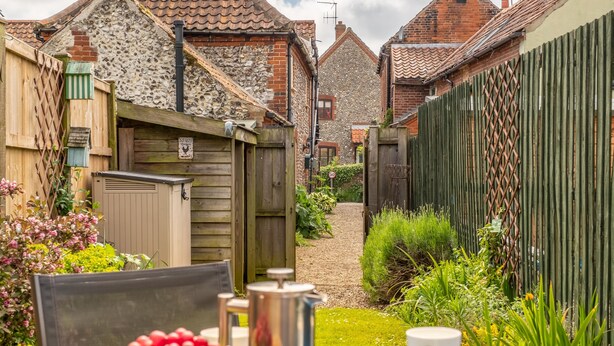 A cozy outdoor seating area with a table set for two, surrounded by a garden with a variety of plants and a wooden fence. - Sherbet Cottage, South Creake, Norfolk (Fakenham)