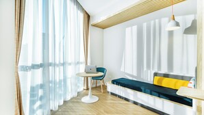 Una habitación moderna con suelo de madera, una mesa redonda con un ordenador portátil, una silla azul, un sofá con cojines amarillos y una lámpara colgante.