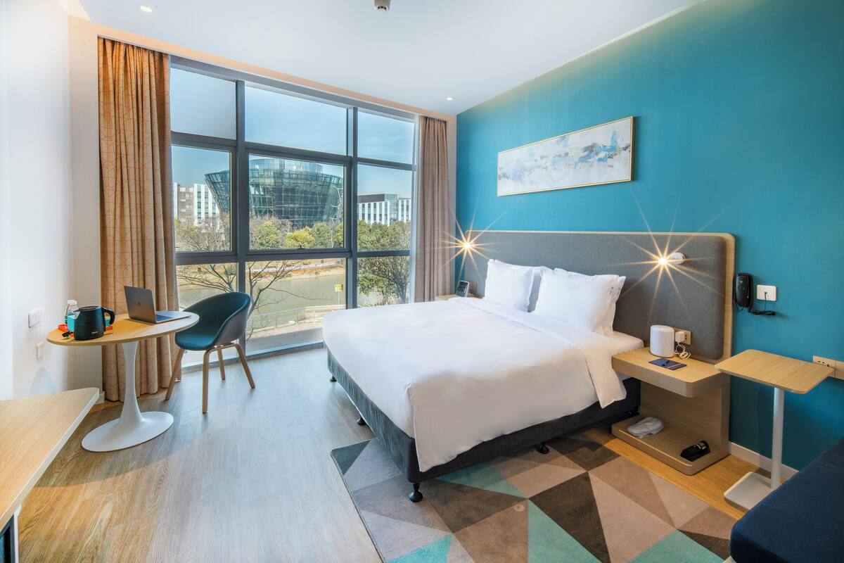 A modern hotel room with a large bed, a desk with a laptop, a chair, and a view of the city through large windows.