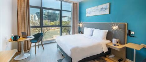 A modern hotel room with a large bed, a desk with a laptop, a chair, and a view of the city through large windows.