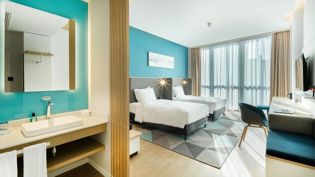 Habitación de hotel moderna con una cama grande, un escritorio, una computadora portátil y un baño con lavamanos y espejo.