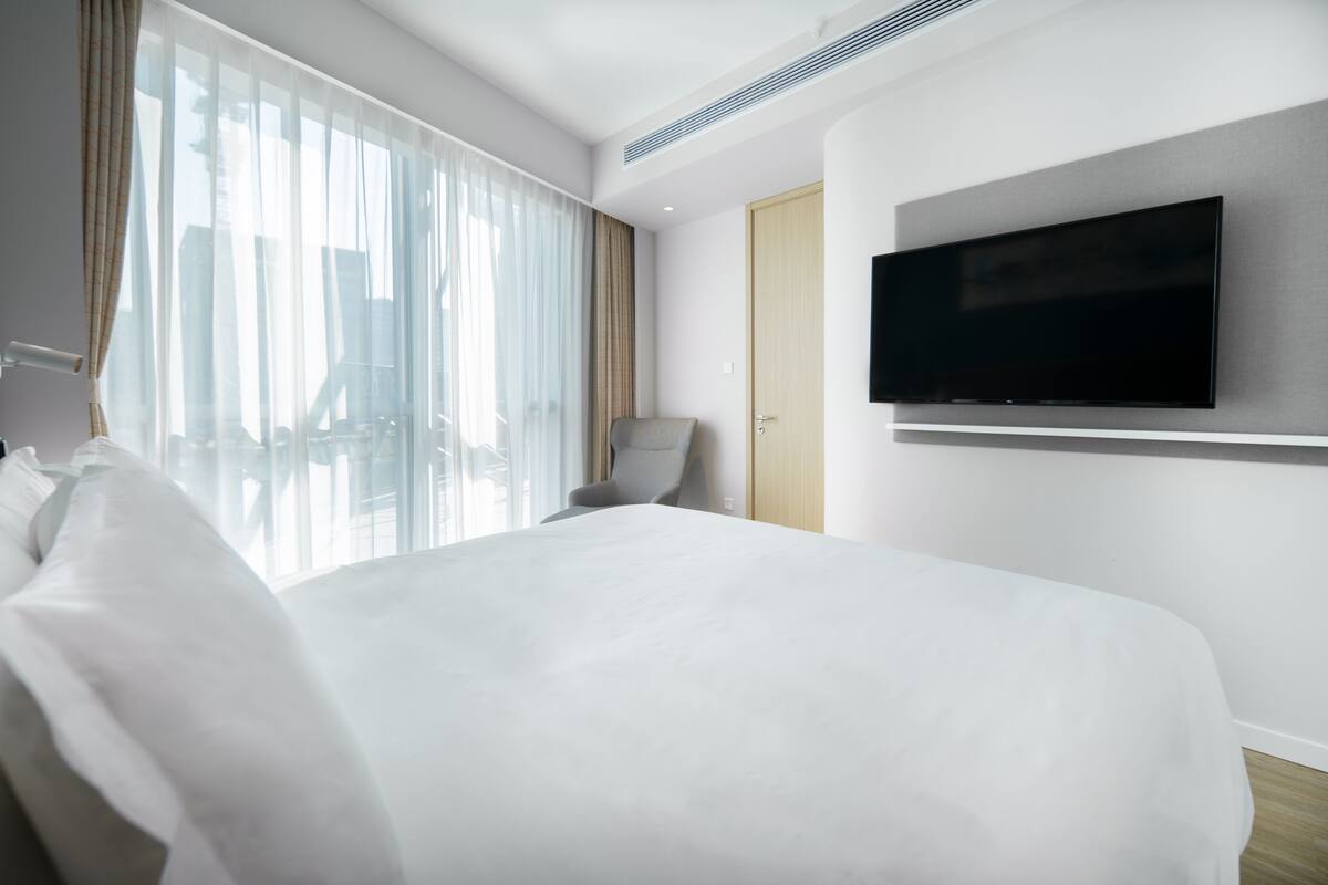 A modern hotel room with a large bed, a flat-screen TV mounted on the wall, a chair, and a large window with sheer curtains.