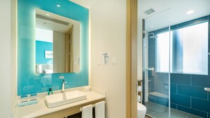 Un baño moderno con un espejo grande, lavamanos blanco y una zona de ducha con mampara de vidrio.