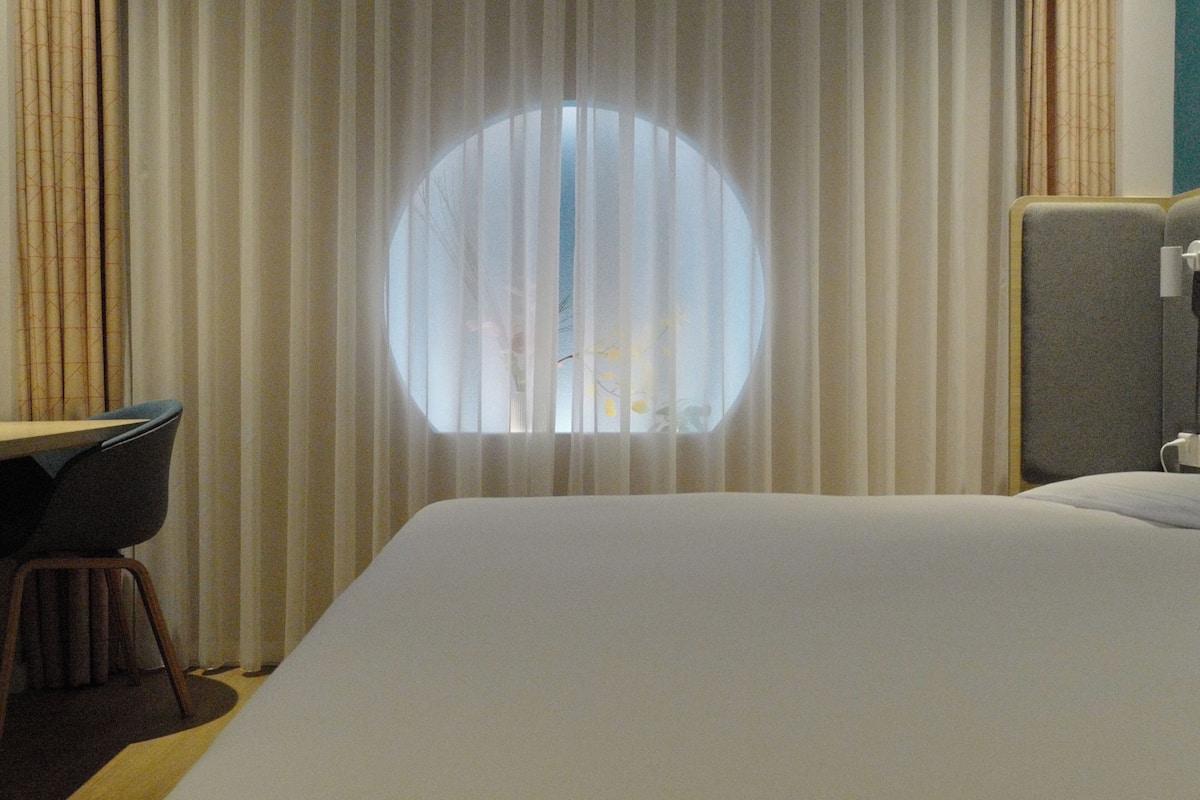 A hotel room with a large bed, a desk, and a chair. There is a window with curtains.