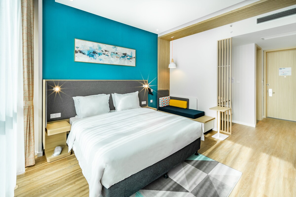A modern hotel room with a large bed, a bedside table, a nightstand, a wall-mounted artwork, and a wooden floor.