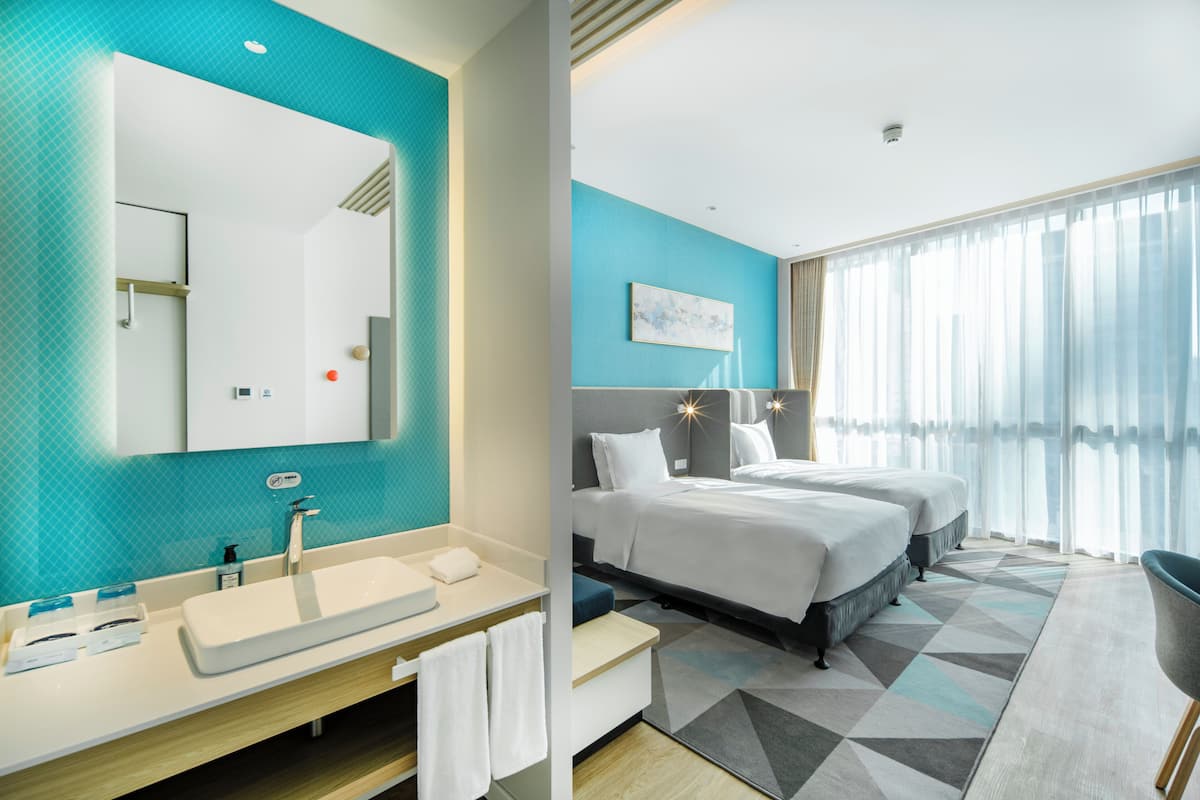 A modern hotel room with a large bed, a separate seating area, a bathroom with a large mirror, and a view of the city through large windows.