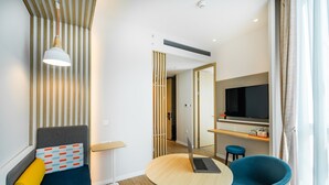 Una habitación de hotel moderna con una cama, una mesa redonda con una computadora portátil, una silla azul, un televisor y un panel de madera.