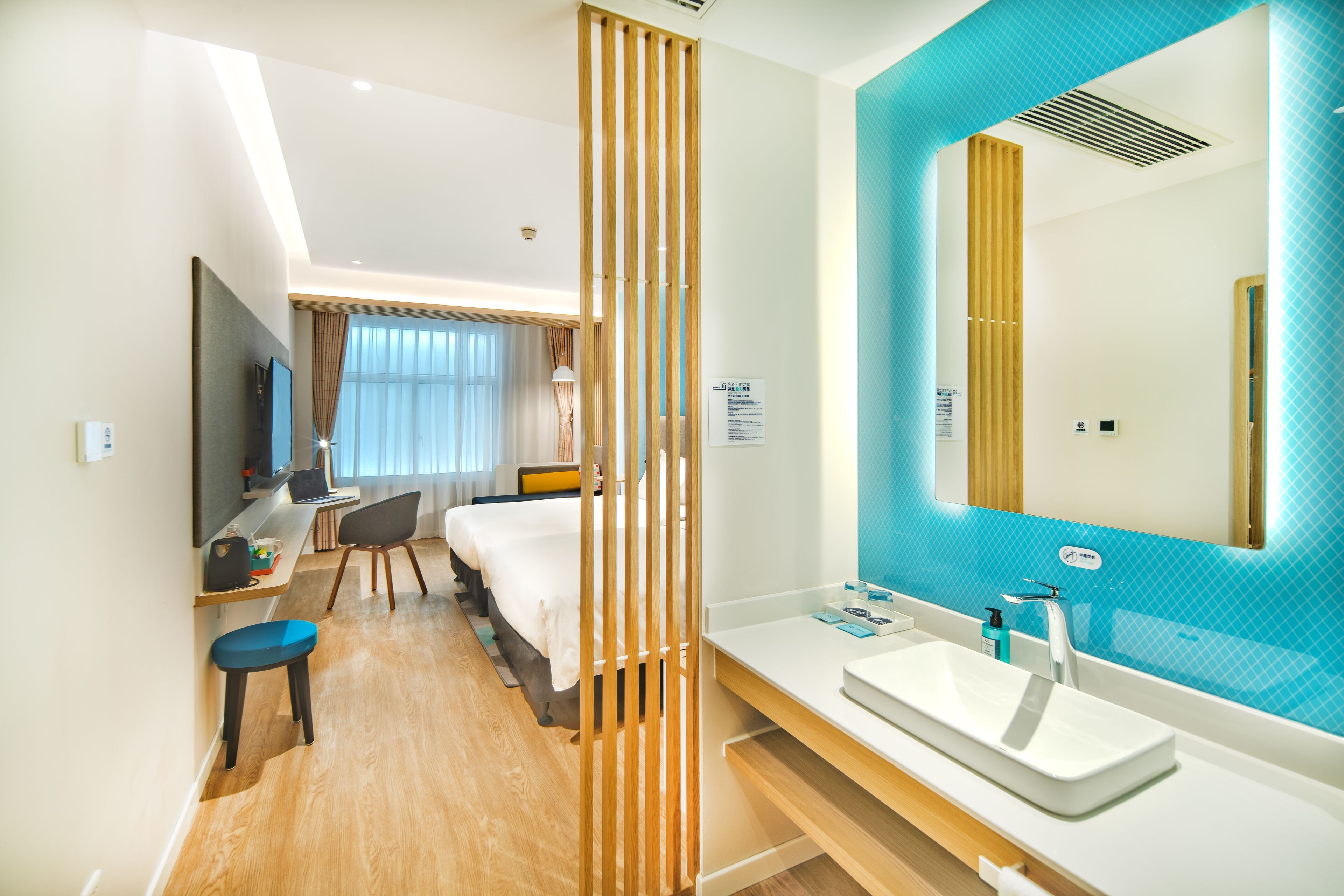 Una habitación de hotel moderna con una cama grande, un escritorio con una computadora portátil, una lámpara de escritorio y un espejo grande con una pared de acento azul.
