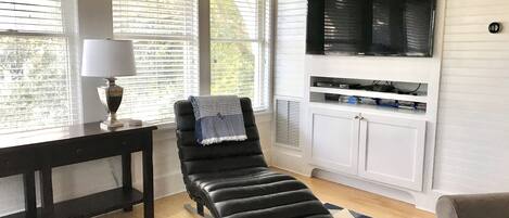 A living room with a black leather sofa, a TV mounted on the wall, a table with a lamp, and a patterned rug.
