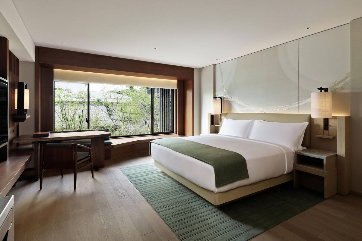 A modern hotel room with a large bed, a desk, and a view of greenery outside.