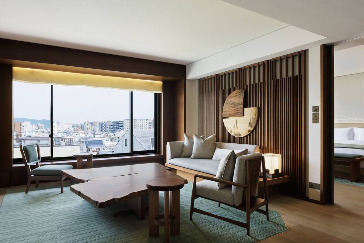 A modern hotel room with a large window, a sofa, a wooden coffee table, and a small side table.