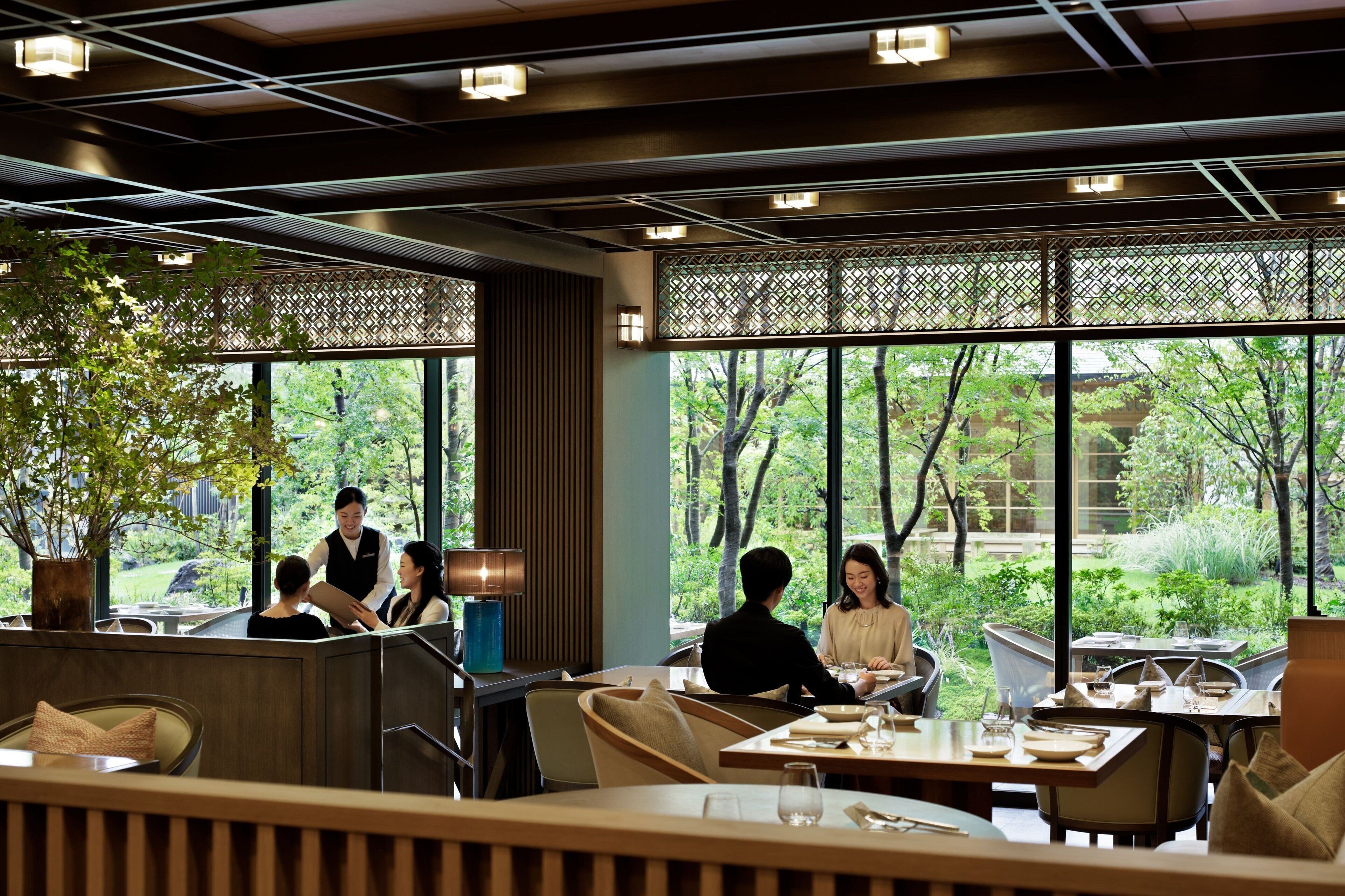 A restaurant with a view of greenery through large windows, wooden paneling, and modern lighting fixtures.