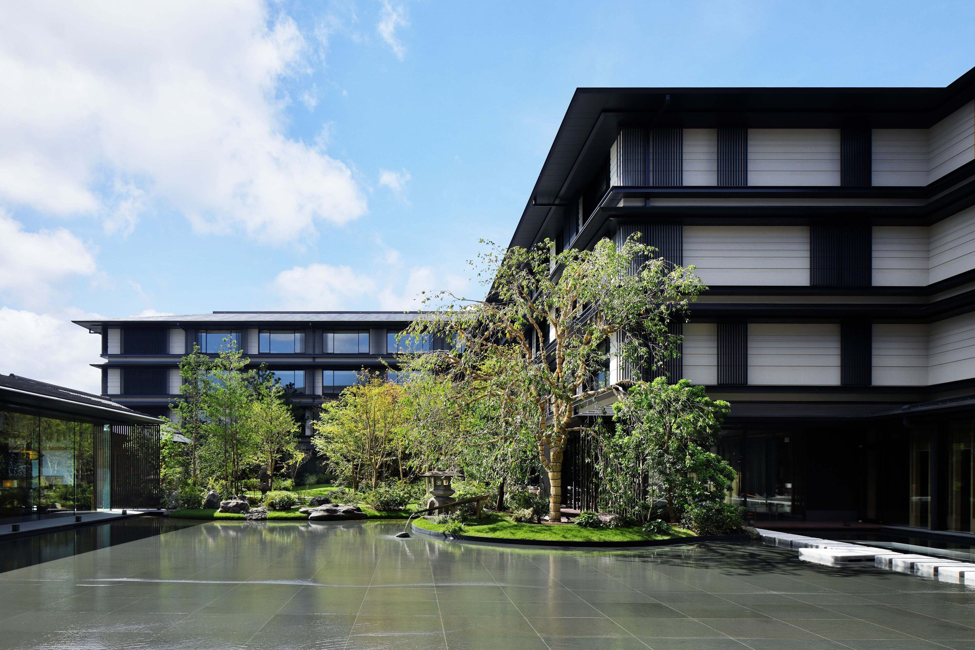 A modern building with a courtyard featuring a reflective water surface, surrounded by greenery and traditional stone lanterns.