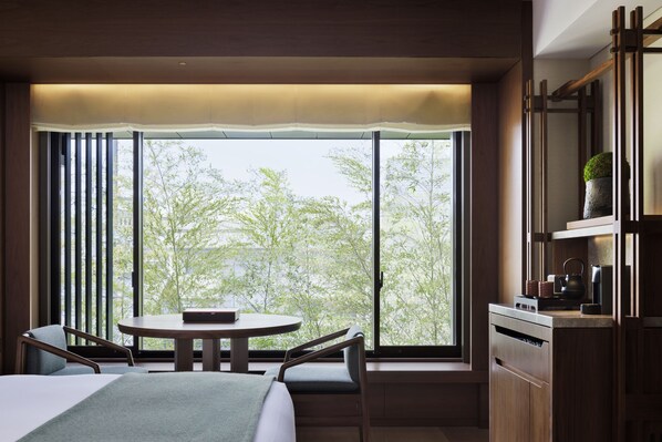 Habitación de hotel con cama, mesa de comedor, dos sillas, un ventanal con vista a árboles y una kitchenette con hervidor y planta.
