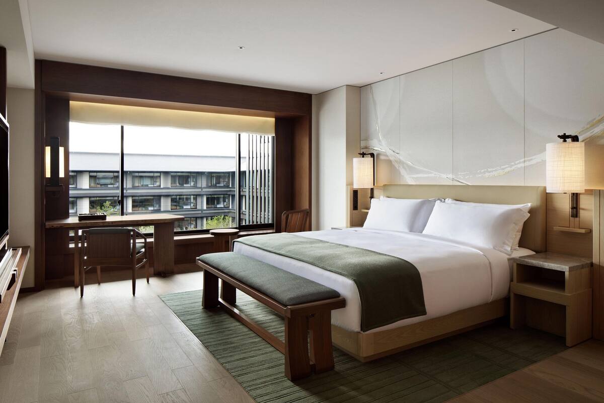 A modern hotel room with a large bed, a desk, and a view of a building through large windows.