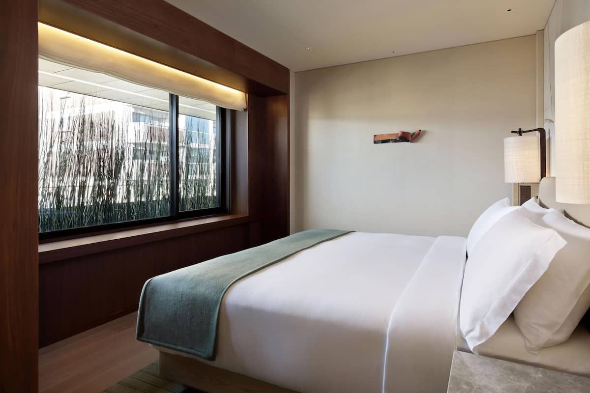 A modern hotel room with a large bed, a window with blinds, a bedside table with a lamp, and a view of trees outside.
