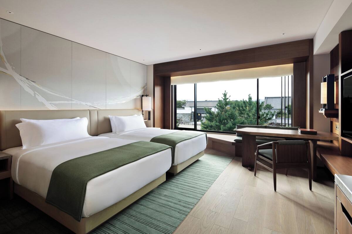 A modern hotel room with two beds, a desk, a chair, and a large window offering a view of the outside.