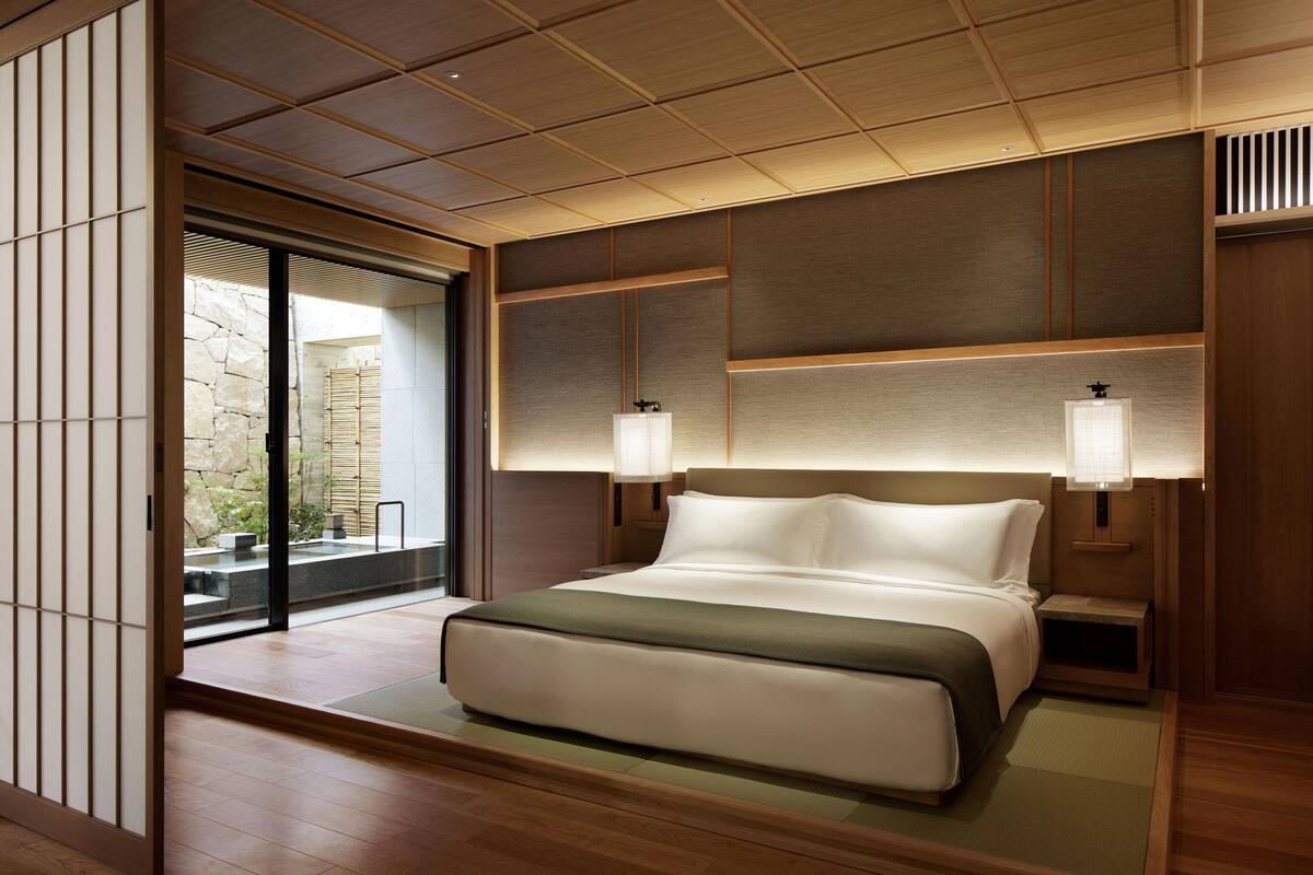 A modern hotel room with a large bed, wooden paneling, and a sliding glass door leading to an outdoor area.