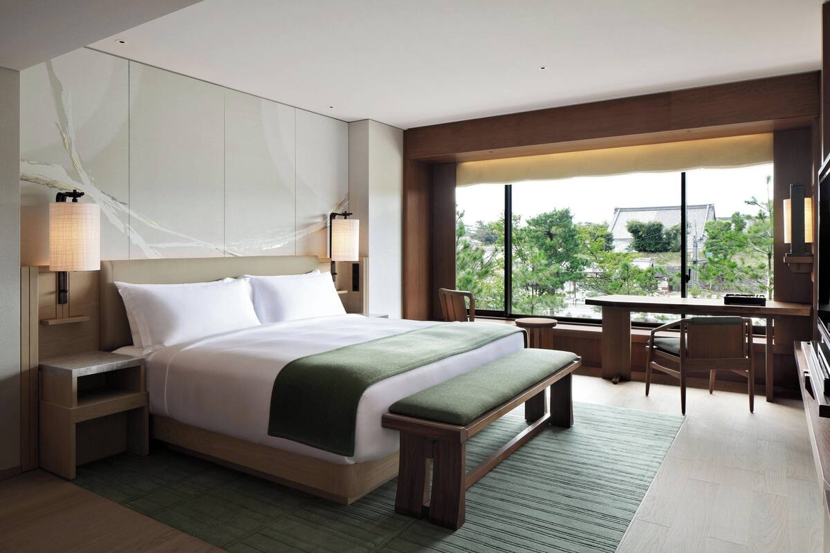 A modern hotel room with a large bed, bedside tables, a desk, and a view of the outdoors.