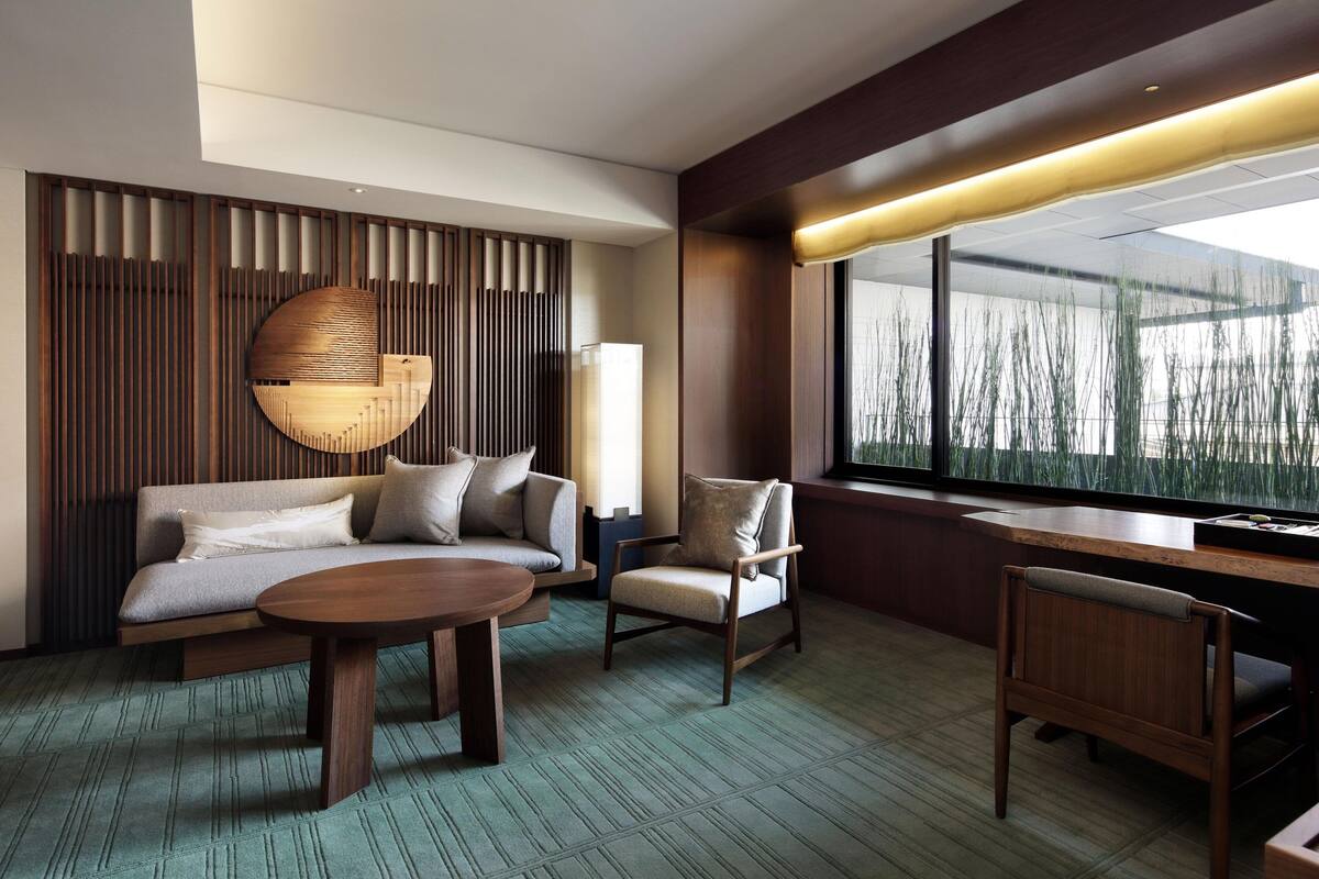 A modern hotel room with a sofa, armchairs, a dining table, and a large window with a view of tall grasses.