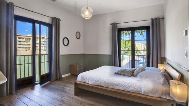 A bedroom with a large bed, a wooden nightstand, a round mirror, and a view of a balcony with a cityscape. - appartement  Serra  : Calvi centre, 300m de la page et du port terrasse bbq (Calvi)