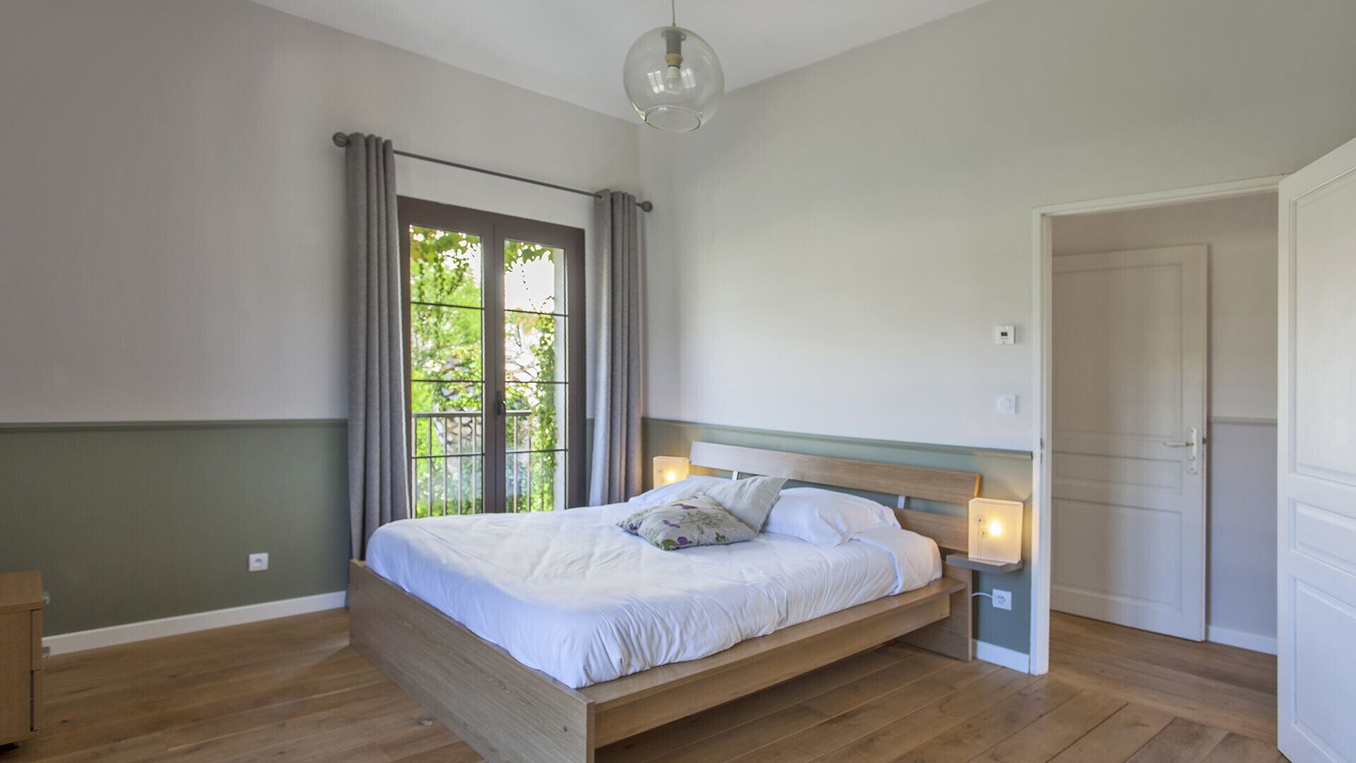 Ein Schlafzimmer mit einem hölzernen Bett, einem Fenster mit Vorhängen, einer Tür und einer Leuchte.